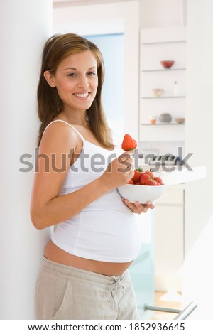 Vertical three quarter profile shot of a young pregnant woman with a bowl of strawberries smiles at the camera.