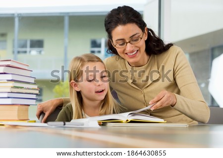 Horizontal shot of a joyous female teacher helping a girl read a book at desk in a classroom.
