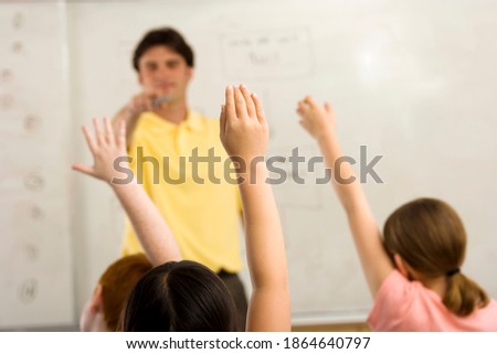 Male teacher pointing at a student with raised hand in a classroom with selective focus on the hand in the foreground.