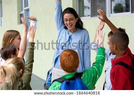 Joyous teacher and students interact outside classroom with raised hands in a corridor.