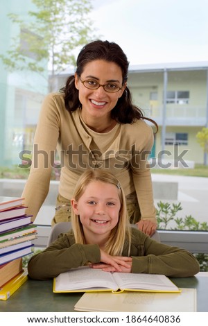 Vertical shot of a seated girl reading a book by her teacher standing behind smile at the camera.