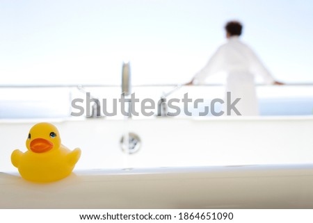 Rubber duck kept on the side of a bath outdoors on a bright, sunny day with a man in a bathrobe standing in the background