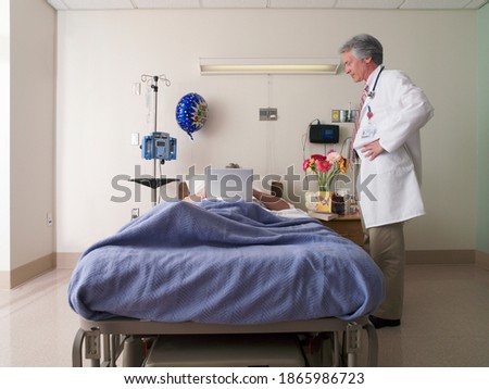 Horizontal shot of a male patient lying in a hospital bed using a laptop with an obscured face as a doctor looks on.