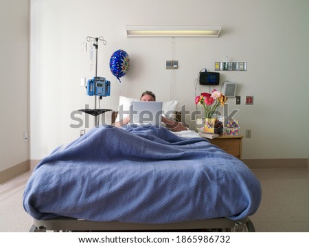 Horizontal shot of a male patient lying in a hospital bed using a laptop with an obscured face.