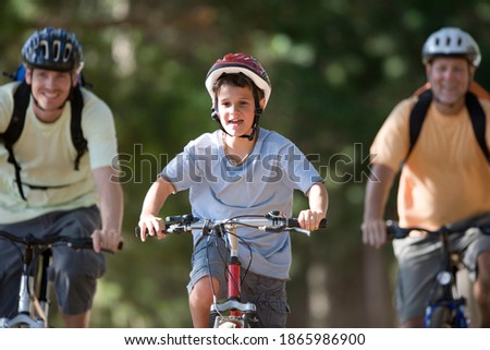 Selective focus on a boy in a cycling helmet mountain biking on a woodland trail with father and grandfather in the background.