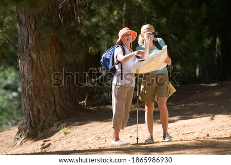 Senior woman and her adult daughter hiking on a woodland trail with the young woman looking through binoculars holding a map.