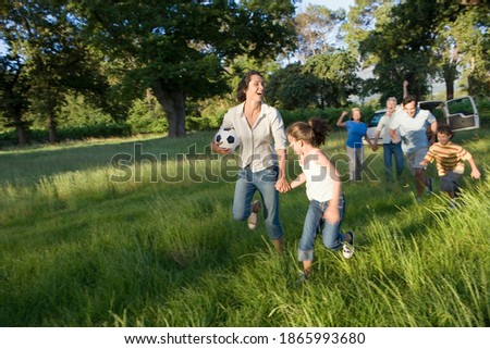 Mother and daughter run together with a soccer ball chased by father and son with grandparents cheering in the background in woodland clearing during camping trip.