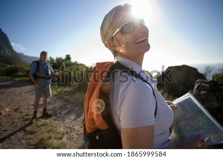 Horizontal waist up profile shot of a smiling mature woman looking at a map on a mountain trail with her husband in the background.