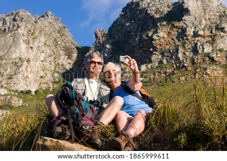 Horizontal low angle shot of a seated mature couple hiking on a mountain trail with the woman taking a self portrait with a camera.
