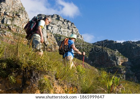 Horizontal low angle profile shot of a mature couple with ruck sacks hiking on a mountain trail on a sunny day.