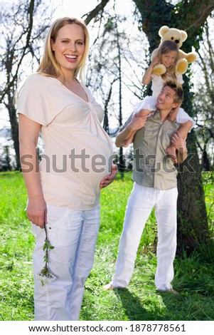 Young pregnant woman with straight blond hair is enjoying a bright, sunny day in the park with her husband and daughter.