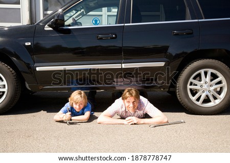 Father and son laying on the ground under a broken down car looking at the camera and smiling as they try to fix it