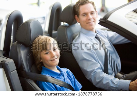 A man is taking his son out for a drive in their new car looking at the camera and smiling.