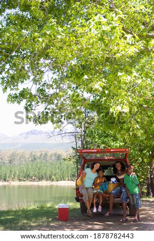 Vertical shot of a family in the trunk of a car under a tree on a sunny day near the lake.