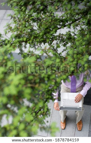 Elevated shot of a businessman working on a laptop on a bench outdoors obscured by a tree.