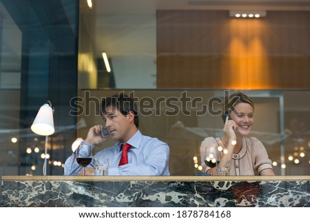 Portrait of a businessman and businesswoman having a drink in the bar and talking on mobile phones seen through a glass window.