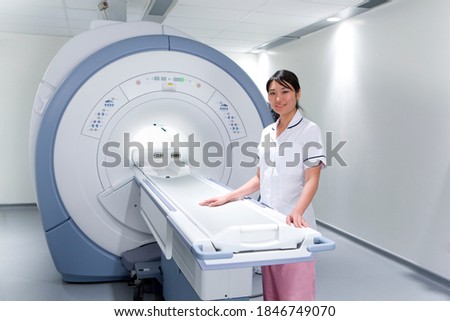 A young adult Radiologist standing next to an MRI scanning machine in a hospital while smiling at the camera
