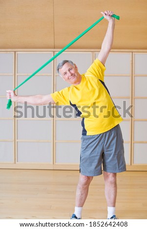 A senior-adult man happily stretching and exercising in a gym using a resistance band while smiling at the camera