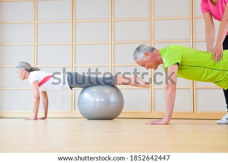 A side view of a gray-haired man and woman balancing their bodies on a fitness ball while being helped and supported by a personal trainer from behind