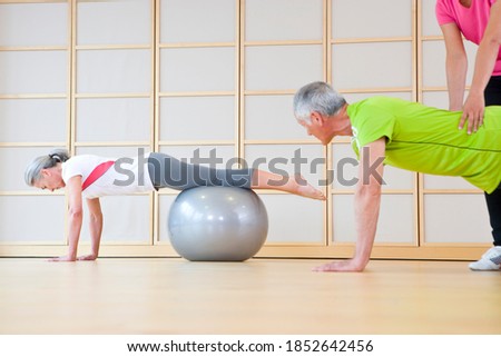 A senior-adult man and woman balancing their bodies on a fitness ball while being helped by their personal trainer from behind