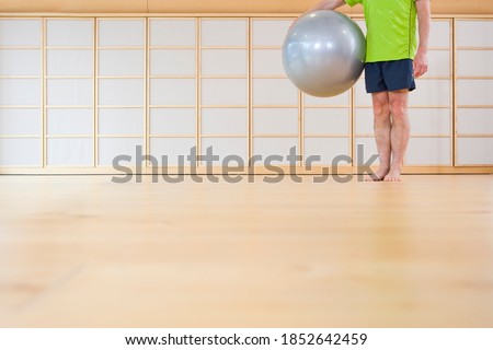 A cropped frontal shot of a senior-adult man standing barefoot in gym while holding a fitness ball