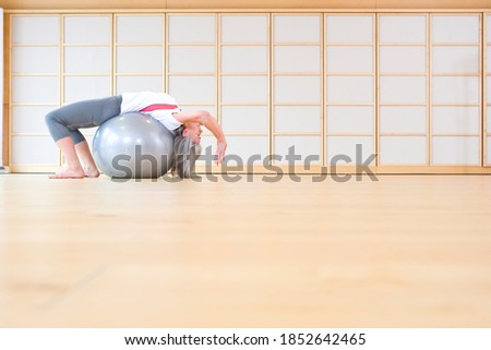 A side view of an elderly woman in gym stretching upside-down on a gray fitness ball