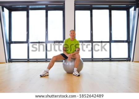 A wide-angle view of an exercising room in a gym with a senior man sitting on fitness ball