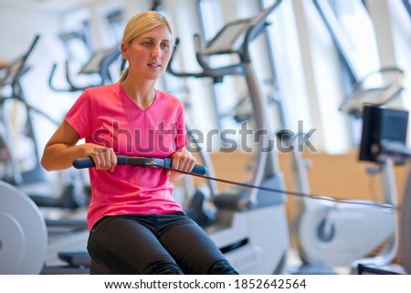 A horizontal portrait of a young-adult woman exercising in the gym on a rowing machine with treadmill in the background