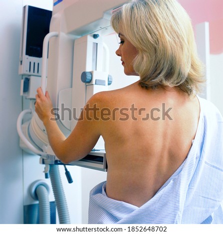 A vertical rear view of a woman in hospital gown taking a mammogram x-ray test in the clinic