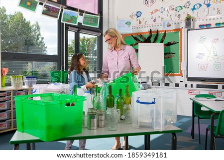 A wide shot of a happy teacher and a young girl sorting recyclable objects in a green tub together in a classroom.