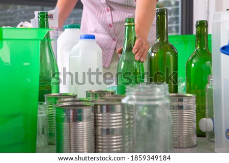 A close up shot of a woman's hand holding a green bottle with other recyclable objects around it.