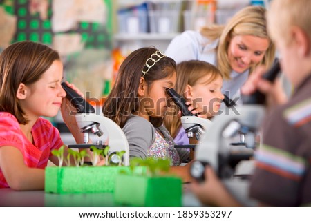 A medium shot of a young girl seriously looking into a microscope in a science class with other students and teacher in background.