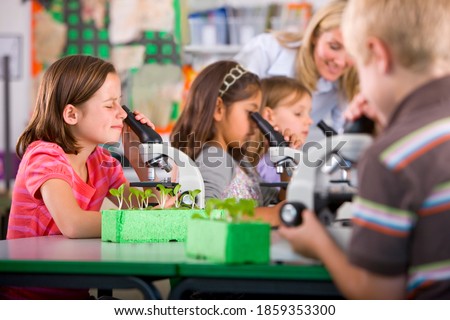 A medium shot of a young girl looking into a microscope in a science class with other students and teacher in background.