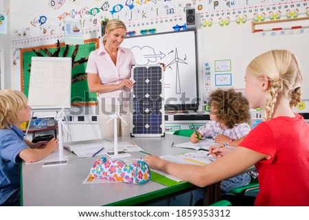 A medium shot of a happy teacher explaining solar panel to her students sitting around a desk with white boards in background.