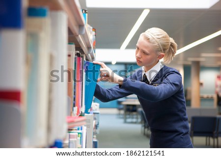 A side profile medium shot of a young blonde girl in school uniform choosing a book from a shelf in a school library