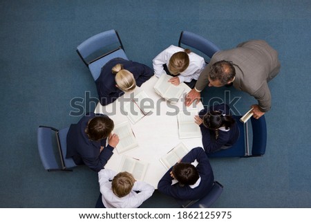 A high angle shot of a teacher guiding his students while they are reading books at a round table.