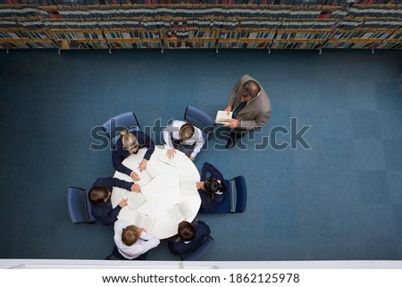 A top angle wide shot of a teacher watching his students reading books at a round table in a library.