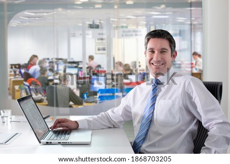 A portrait of a smiling businessman sitting at his desk using a laptop in an office with people working in the background