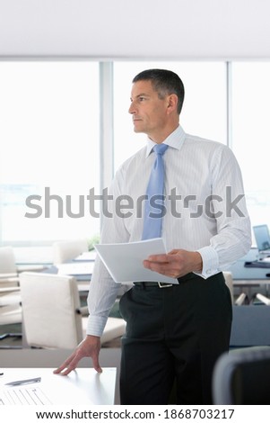 A pensive businessman in formal wear standing next to a desk in an office while holding important paperwork