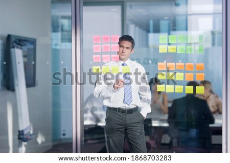 A pensive businessman closely looking at the adhesive notes on the glass wall while co-workers having a meeting in the background