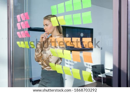 A view of a pensive businesswoman through a glass wall closely looking at the adhesive notes in an office