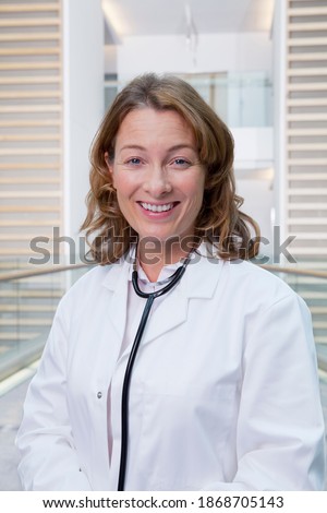 A vertical portrait of a smiling doctor in a lab coat while standing in the hospital corridor with a stethoscope around her neck