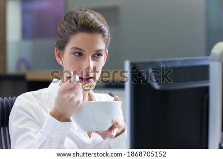 A businesswoman eating cereal from a bowl at her desk while working on a computer in the office