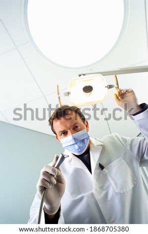 A low angle portrait of a dentist with a lab coat and a surgical mask holding the drill while standing under a lamp