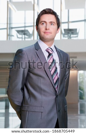 A vertical view of a serious businessman in a formal suit standing in the office building with his hands clasped behind