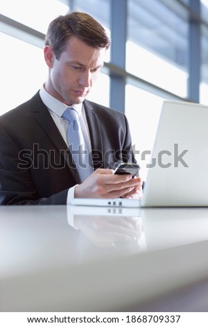 A vertical view of a Businessman in formal suit using a laptop and a cell phone for text messaging at a table in the office