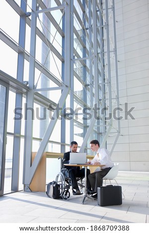 A full-frame vertical view of a businessman in wheelchair using laptop with his co-worker while sitting at a table next to their suitcases in a modern office