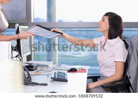A side view of a businesswoman handing over a report to her co-worker while sitting at her desk in the office