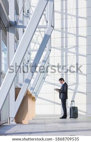 A full-length vertical view of a businessman with suitcase using a digital tablet while standing in the modern lobby