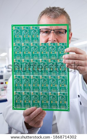 A vertical closeup of an engineer in selective focus examining a printed circuit board while looking through its gaps in a laboratory